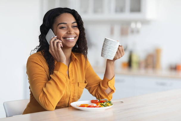 Rahat Afrikalı Amerikalı kadın kahvaltı yapıyor, telefonda konuşuyor. - Fotoğraf, Görsel