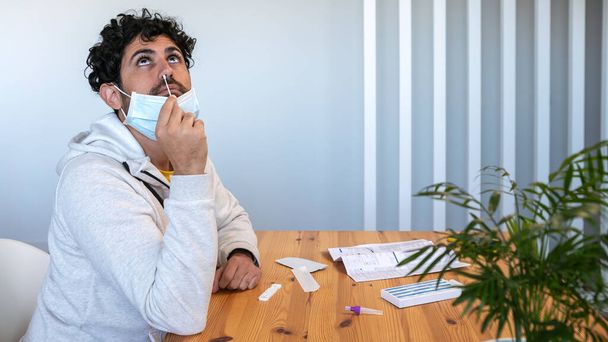 Un uomo caucasico che indossa una maschera inserisce un batuffolo di cotone in un naso per raccogliere un possibile campione positivo di COVID-19 durante la pandemia. Procedura di prova dell'antigene a casa. - Foto, immagini