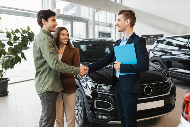 Heureux jeune couple caucasien acheter une nouvelle voiture, serrer la main avec un vendeur confiant chez le concessionnaire - Photo, image
