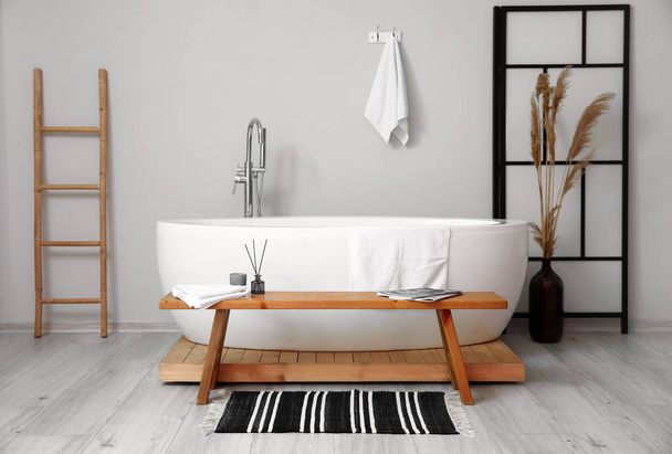 Интерьер светлой комнаты с белой ванной и деревянной скамейкой - Фото, изображение