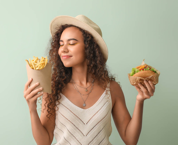 Young African-American woman with french fries and burger on color background - Photo, Image