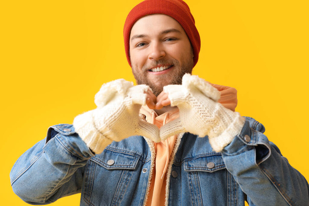 Young man making heart shape with his hands in warm gloves on yellow background - Photo, Image