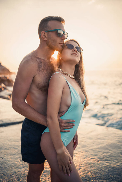 Um casal amoroso caminha ao longo da praia à beira-mar. Família jovem ao pôr-do-sol junto ao Mar Mediterrâneo. Conceito de férias. Uma mulher de fato de banho e um homem de calções ao pôr-do-sol junto ao mar. Foco seletivo. - Foto, Imagem