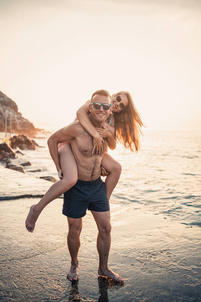 Egy szerető pár sétál a tengerparton. Fiatal család naplementekor a Földközi-tengernél. Szabadság koncepció. Egy nő fürdőruhában, és egy férfi rövidnadrágban naplementekor a tengernél. Szelektív fókusz. - Fotó, kép