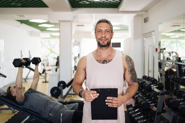 Persönlicher Fitnesstrainer schreibt Trainingsplan in der Nähe seines Trainees auf Fitnessstudio-Hintergrund - Foto, Bild