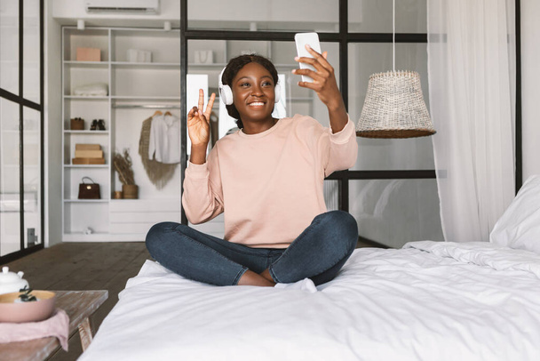 African Lady Making Selfie On Cellphone Gesturing Victory In Bedroom - 写真・画像