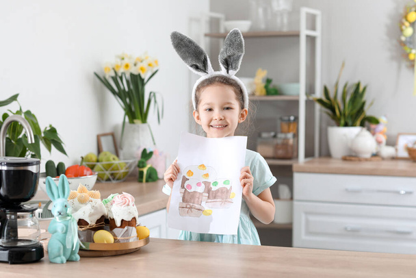 Χαριτωμένο κοριτσάκι με αυτιά κουνελιού και ευχετήρια κάρτα Πάσχα στην κουζίνα στο σπίτι - Φωτογραφία, εικόνα