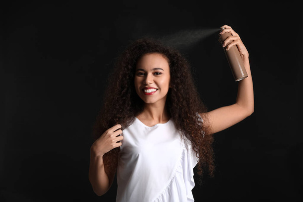 Mujer afroamericana bastante joven con spray para el cabello sobre fondo negro - Foto, Imagen