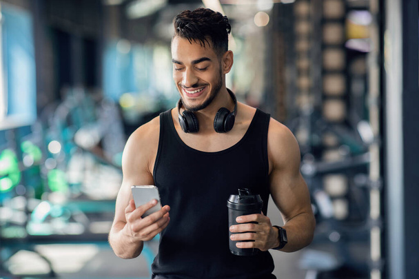 Lächelnder arabischer Sportler entspannt sich nach dem Training im Fitnessstudio mit Smartphone - Foto, Bild