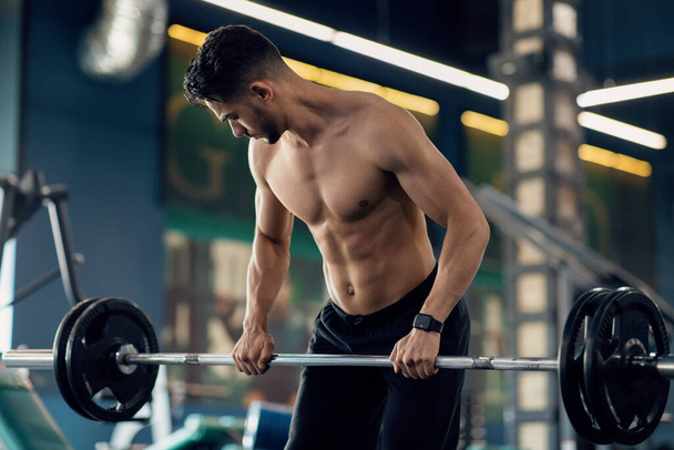 hombre árabe musculoso sin camisa levantando pesadas barras mientras se entrena en el gimnasio - Foto, Imagen