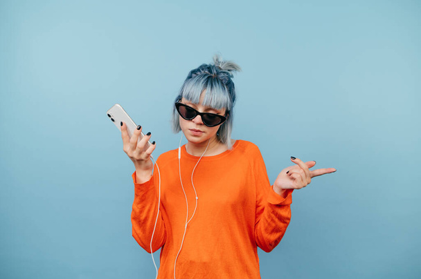 Fényes hipster lány napszemüvegben okostelefonnal a kezében, és a fejhallgató táncol egy komoly arc, és zenét hallgat a kék háttér. - Fotó, kép