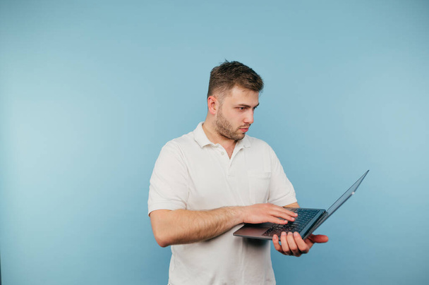 Hombre adulto en una camiseta blanca se para con una cara seria sobre un fondo azul y trabaja en una computadora portátil - Foto, Imagen