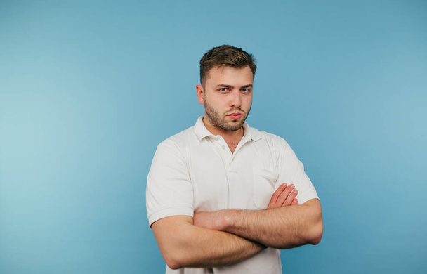 Homem barbudo sério em uma camiseta branca isolada em um fundo azul, olhando atentamente para a câmera. - Foto, Imagem