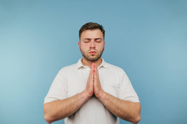 Calm adult man in white t-shirt praying with closed eyes on blue background. - Fotoğraf, Görsel