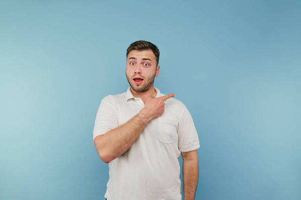 Shocked young man in a white T-shirt with bristles points his finger to the side with a surprised face on a blue background. - Photo, Image