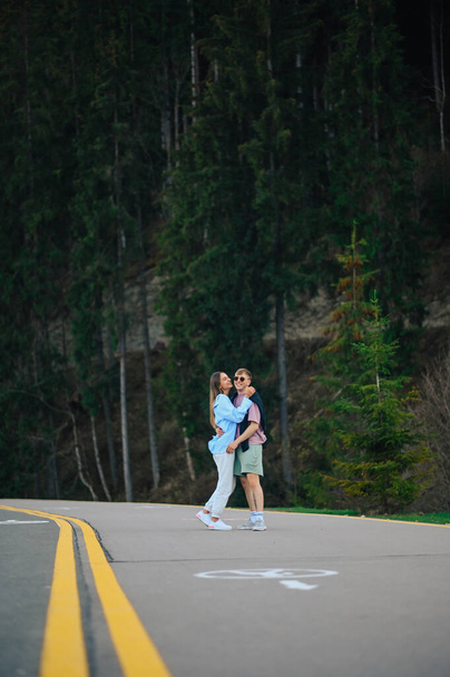Όμορφο ζευγάρι αγκαλιάζει σε ένα θέρετρο στα βουνά σε μια βόλτα, στέκεται στο δρόμο με φόντο το δάσος. - Φωτογραφία, εικόνα