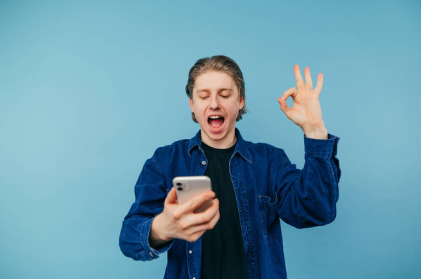 Portrait of a joyful guy in a blue shirt on a blue background with a smartphone in his hands enjoys and shows a gesture of ok. - Photo, Image
