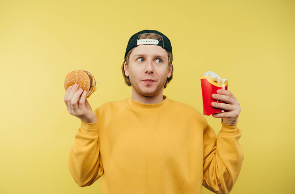 Vicces fickó sapkában, sárga pulóverben, sárga háttérrel, sült krumplival és hamburgerrel a kezében. Fiatalember és gyorskaja. - Fotó, kép