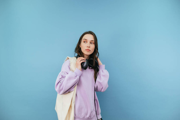 Attractive woman in casual clothes and headphones with a shopper bag in her hands isolated on a blue background, looking away with a serious face - Photo, image
