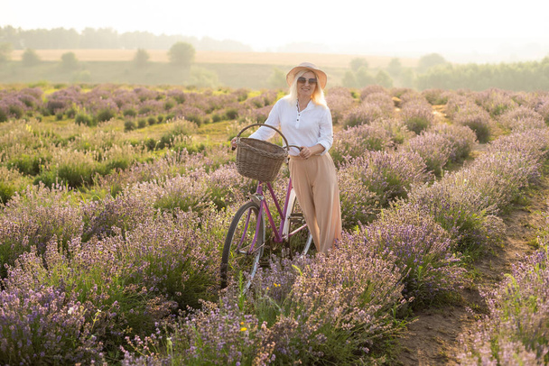Fiatal szőke utazó szalmakalapot visel a levendula mezőn, levendula virágokkal körülvéve.. - Fotó, kép