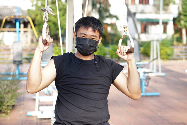 屋外公園で黒顔のマスクをしたアジア人男性が運動マシンで運動している。概念:ヘルスケア、コロナウイルスまたはCovid-19からの自己保護。公園を利用する際の自己認識.         - 写真・画像