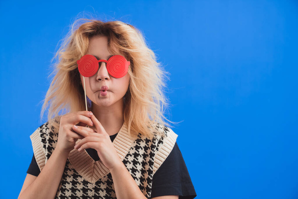 Смішна блондинка-волохата кавказька дівчина в 20 років робить каченя і прикриває очі підробленими червоними паперовими окулярами на синьому фоні
. - Фото, зображення