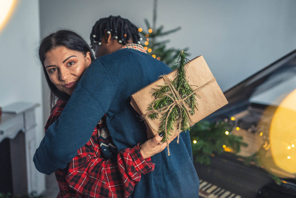 Noel 'i kutlayan ırklar arası bir çift. İki eş arasında kucaklaşın. Beyaz kadın kameraya hediyesini gösteriyor.. - Fotoğraf, Görsel