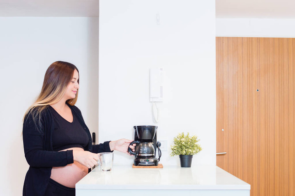 jovem mulher branca grávida na cozinha de seu apartamento preparando café da manhã e café para o marido, tomando a panela de café da panela no balcão da cozinha. - Foto, Imagem