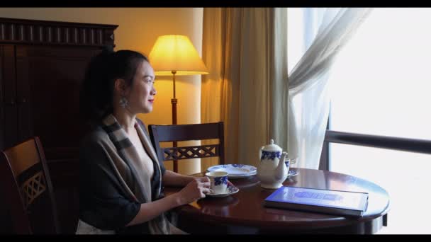 Asiático origem meia idade mulher atraente sentado em retro vintage sala mesa cadeira armário chão lâmpada chá copo conjunto janela luz trabalho a partir de casa sentado olhando para fora pensando  - Filmagem, Vídeo