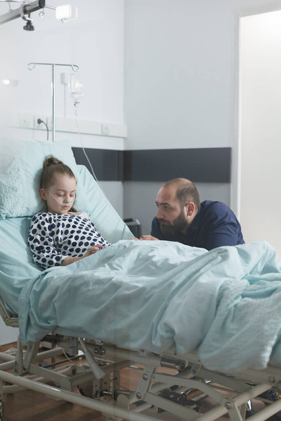 Hombre triste ansioso viendo a su hijita enferma durmiendo en la cama de la clínica mientras está en el hospital pediátrico. - Foto, Imagen