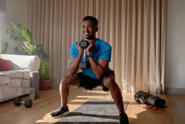 African American male squatting in lounge holding dumbbell weight, working out at home - Zdjęcie, obraz