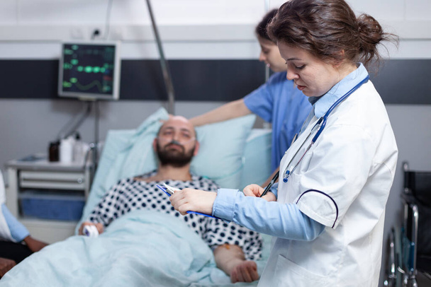 Doktor se stetoskopem čtení lékařské karty, zatímco zdravotní sestra dělá pacienta pohodlné - Fotografie, Obrázek