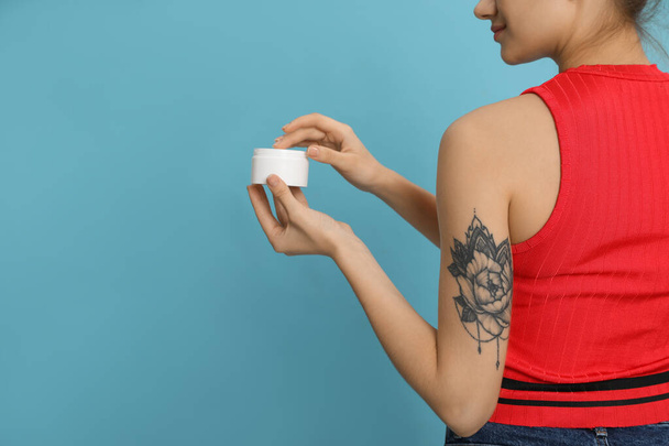 Woman with tattoo holding jar of cream against light blue background, closeup. Space for text - Fotografie, Obrázek
