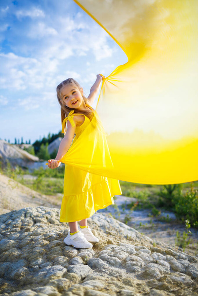 湖によって黄色の生地に翼を持つ黄色のドレスの小さな6 〜 7歳のウクライナの少女。青い空と黄色の布、ウクライナの国旗の色. - 写真・画像