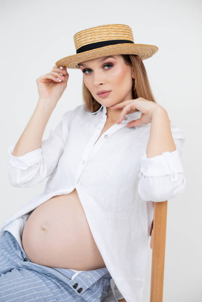Portret van jonge zwangere vrouw met lang donker haar en make-up dragen wit shirt, open gestreepte jeans en strohoed. - Foto, afbeelding