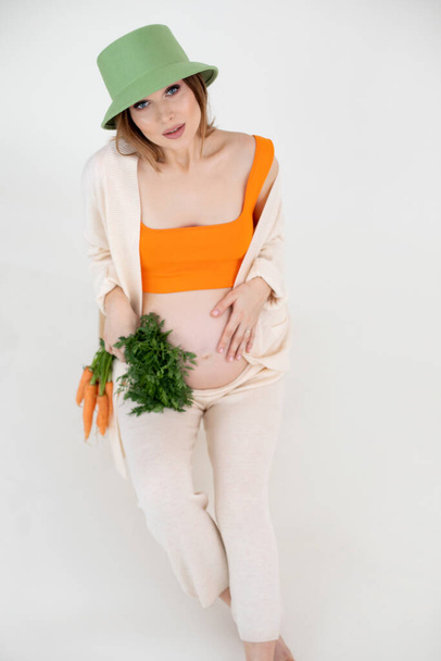 Young gorgeous pregnant woman with long dark hair wear green bucket hat, sitting, holding belly and bunch of carrots. - Photo, Image