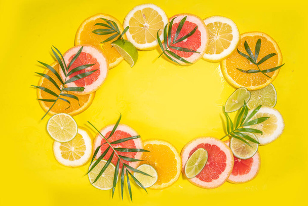 柑橘類の果実スライスと熱帯ヤシの葉のカラフルなフラットレイ。オレンジ、グレープフルーツ、レモン、ライムの黄色の背景に明るい色のスライス。夏休みの背景｜Top view - 写真・画像