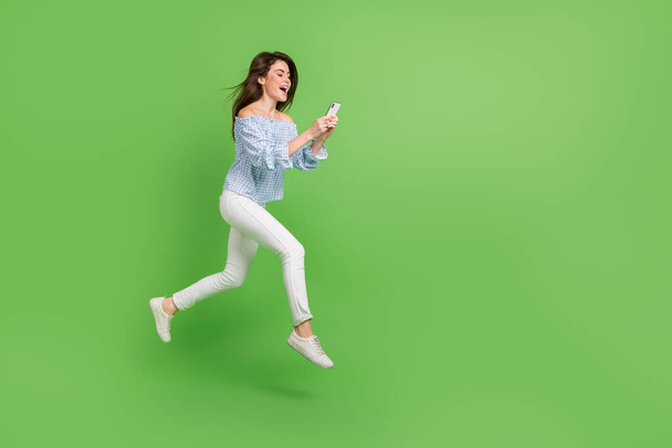 Photo of excited woman wear plaid off-shoulders blouse jumping high chatting gadget empty space isolated green color background - Zdjęcie, obraz