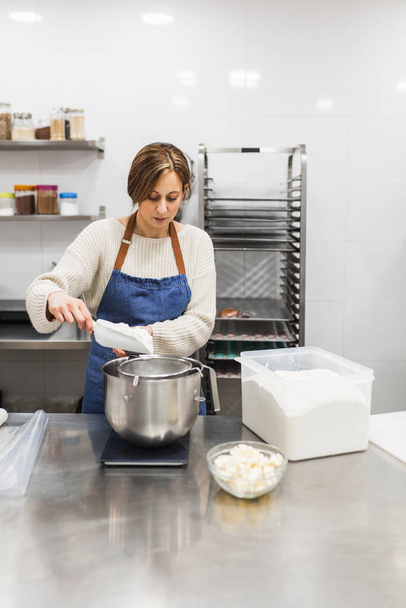 Kobieta cukiernik w fartuchu wylewanie mąki do miski podczas gotowania w kuchni. - Zdjęcie, obraz