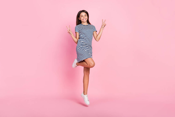 Photo of girly sweet girl dressed striped dress jumping high showing v-signs isolated pink color background - Foto, immagini
