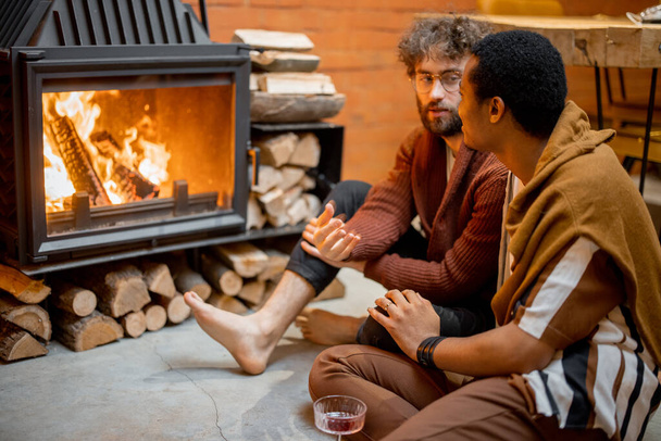 Two men sitting together by the burning fireplace at cozy home - Фото, изображение