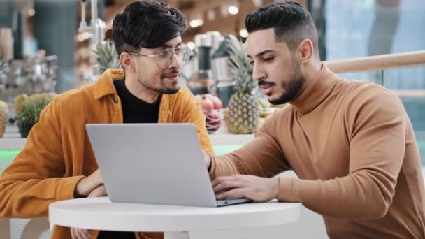 Deux hommes arabes assis regardant l'écran d'ordinateur portable communiquer sourires étudier en utilisant de nouvelles applications informatiques pages web de navigation en ligne sortir ensemble mise en place de logiciels jeune gars expliquant à un ami aider à soutenir - Séquence, vidéo