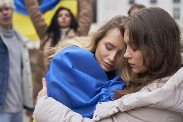 Zwei junge kaukasische Frauen umarmen sich vor und im Hintergrund eine Gruppe von Menschen, die gegen den Krieg in der Ukraine demonstrieren - Foto, Bild