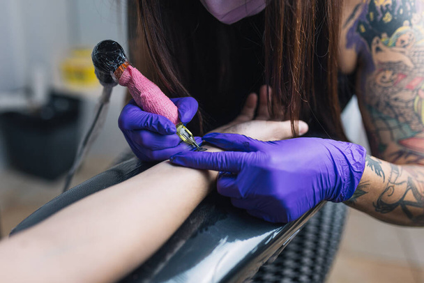 Close-up van de tattoo machine. Een vrouwelijke tattoo meester in een masker en handschoenen creëert een patroon op de huid van een vrouw. - Foto, afbeelding
