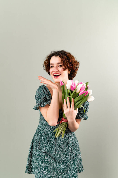 Удивлённая девочка-подросток с кудряшками на сером фоне с улыбающимися тюльпанами. В летнем платье. Копирование пространства - Фото, изображение
