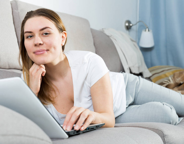 Cheerful girl is lying on sofa and playful posing with laptop - Foto, Imagem