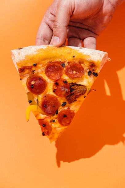 Stuk pizza in de hand met chorizo worst, paprika 's, zongedroogde tomaten, gedroogde chili pepers, gouda kaas en mozzarella kaas met pilatensaus, op een dunne korst en op een pluizig bord. oranje achtergrond. copyright. bovenaanzicht - Foto, afbeelding