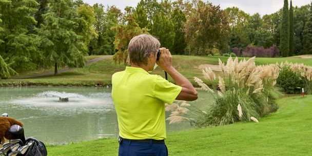 ゴルファーはレーザーレンジファインダーでゴルフコースの旗までの距離を正確に測定します。背景には森. - 写真・画像