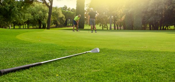 Golf sopası elinde golf sopasıyla topu deliğe sokmaya çalışıyor. Yeşildeki bir oyuncu, topu bayrağa doğrultmadan önce eğimleri ve delikten uzaklığı ölçer.. - Fotoğraf, Görsel