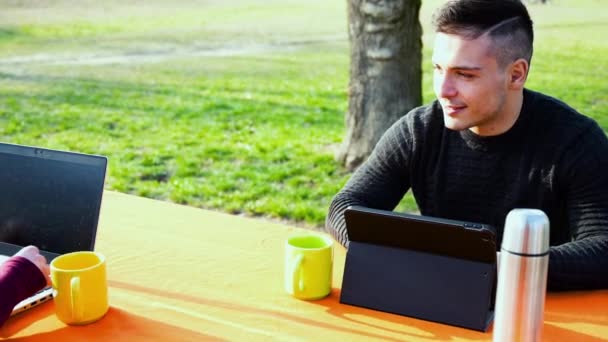 Una pareja joven trabaja remotamente en un parque en un día soleado agradable. Utilizan un ordenador portátil y una tableta mientras que hablan entre sí. En la mesa hay tazas y un flask.Smiling, gente independiente que usa tecnología - Imágenes, Vídeo
